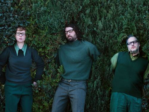 Eine Frau und zwei Männer, alle mit Brille, drücken sich in eine grüne Hecke.